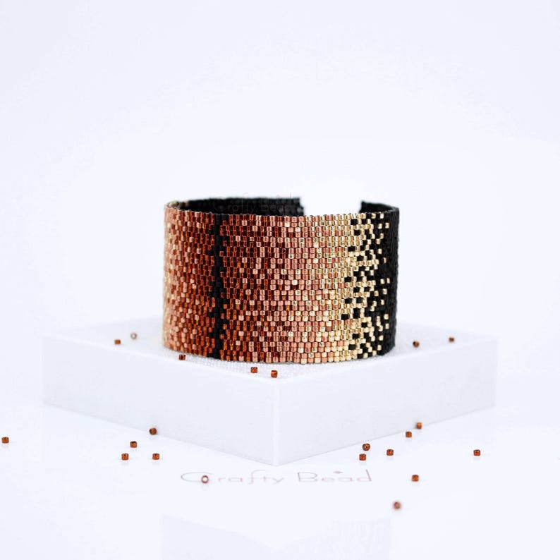 Op de afbeelding: Een zwarte, koperen en gouden kralen armband met een geometrisch patroon. De armband ligt op een wit oppervlak met kleine koperen kralen eromheen.