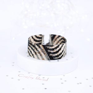 K&ouml;nnte beinhalten: Ein Armband aus schwarzen, wei&szlig;en und goldenen Perlen mit Zebra-Muster. Das Armband befindet sich auf einem wei&szlig;en Pr&auml;sentationsst&auml;nder mit dem aufgedruckten Text "Crafty Bead".