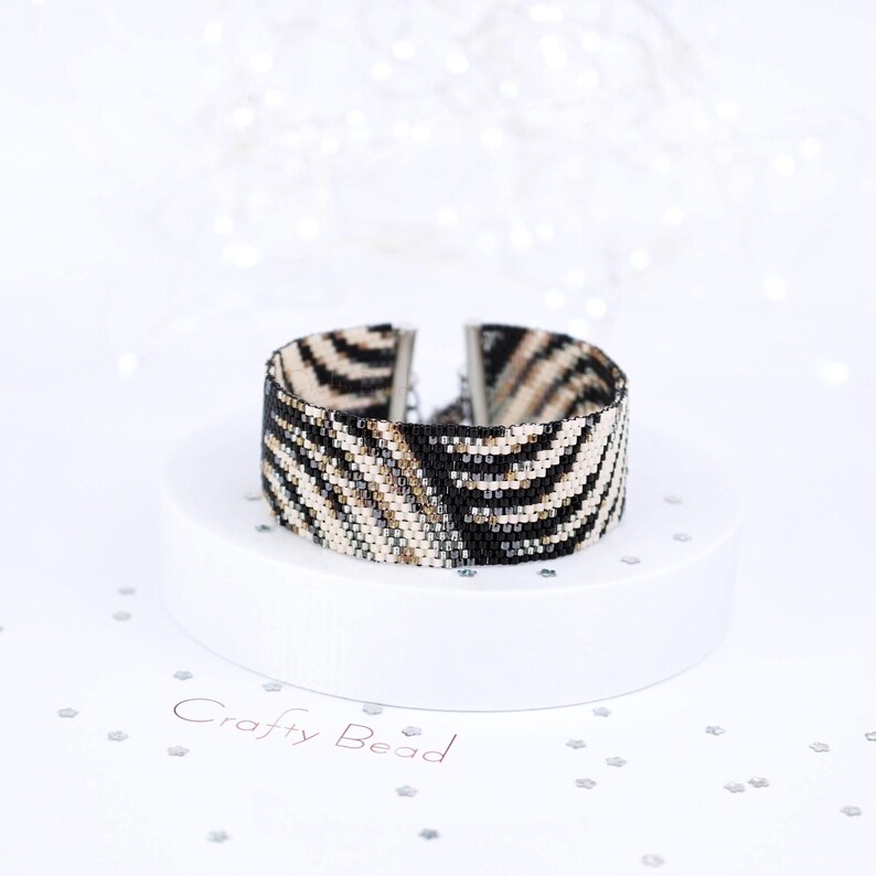 K&ouml;nnte beinhalten: Ein Armband aus schwarzen, wei&szlig;en und goldenen Perlen mit Zebra-Muster. Das Armband befindet sich auf einem wei&szlig;en Pr&auml;sentationsst&auml;nder mit dem aufgedruckten Text "Crafty Bead".
