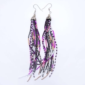 Pode incluir: Um par de brincos longos de contas com uma parte superior prateada. As contas são em tons de roxo, rosa, branco e preto.