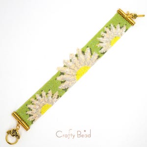 Pu&ograve; includere: Un braccialetto di perline verdi con fiori di margherita bianchi e chiusure dorate. Il braccialetto &egrave; realizzato con perline a seme e presenta un delicato motivo floreale. "Crafty Bead" &egrave; stampato sullo sfondo bianco.