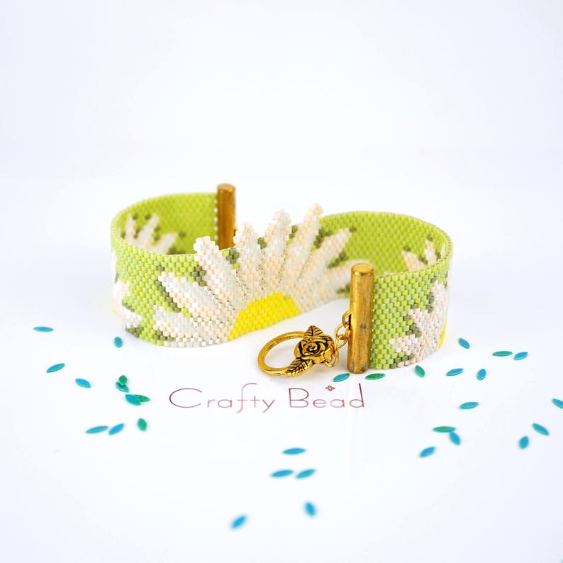 Pu&ograve; includere: Un braccialetto di perline verde e bianco con un design a margherita e una chiusura dorata. Il braccialetto &egrave; su una superficie bianca con coriandoli blu e verdi.