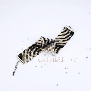 K&ouml;nnte beinhalten: Ein schwarz-wei&szlig;es Armband mit Zebra-Muster und silbernen Verschl&uuml;ssen. Das Armband ist zu einem Knoten gebunden und auf einer wei&szlig;en Oberfl&auml;che mit silbernem Glitzer fotografiert.