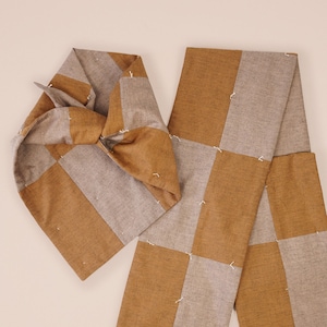 Może przedstawiać: Szalik z tkaniny w kratę w kolorze brązowym i szarym z zawiązanym końcem. Szalik jest wykonany z miękkiego, tkanego materiału i ma subtelną fakturę.