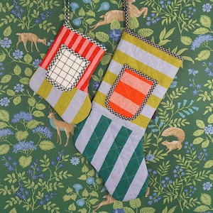 Puede incluir: Dos calcetines de Navidad a rayas con un esquema de color verde, azul y blanco. Los calcetines tienen un bolsillo en la parte delantera con un color y un patrón contrastantes. Los calcetines están colgados sobre un fondo verde con un patrón floral.