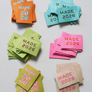May include: Stacks of fabric labels in various colors, including orange, teal, green, pink, and beige. Each label has the text "MADE IN 2026" in a contrasting color. The labels are neatly arranged, showcasing the text prominently.