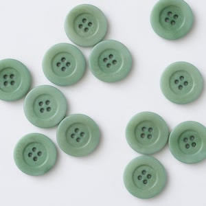 Puede incluir: Un conjunto de botones redondos verde salvia, cada uno con cuatro agujeros. Los botones están dispuestos sobre una superficie blanca, mostrando su tamaño y color uniformes. Ideales para proyectos de costura y manualidades.