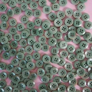 Puede incluir: Un conjunto de botones redondos verde salvia con cuatro agujeros cada uno, dispersos sobre una superficie rosa. Los botones tienen un aspecto ligeramente moteado. La imagen muestra una colección de botones, ideal para coser o manualidades.