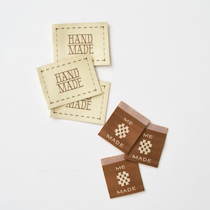 May include: A collection of rectangular fabric labels. Three cream-colored labels feature a dashed border and the text "HAND MADE" in brown. Three brown labels feature the text "ME" above a small woven square icon and "MADE" below.