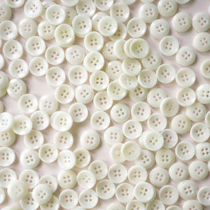 May include: A collection of ivory-colored, round buttons with four holes each. The buttons are scattered across a surface, creating a textured pattern. The buttons are made of a smooth, opaque material.
