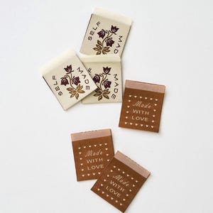 May include: A collection of fabric labels including three cream-colored tags with a dark brown floral design and the text SELF MADE, and three brown tags featuring the text Made WITH LOVE surrounded by a border of small white hearts.