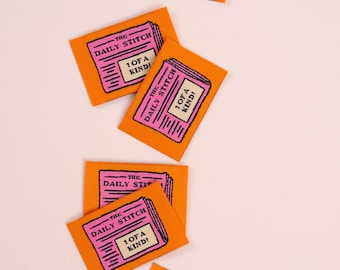 May include: Several orange fabric labels featuring a pink newspaper design. The labels display the text "THE DAILY STITCH" and "1 OF A KIND!" These are likely for craft projects.