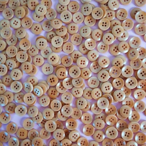Può includere: Una collezione di bottoni in legno marrone chiaro, ciascuno con quattro fori, sparsi su una superficie rosa pallido. I bottoni presentano variazioni di colore e venature, creando un effetto strutturato. Ogni bottone misura circa 2 cm di diametro.