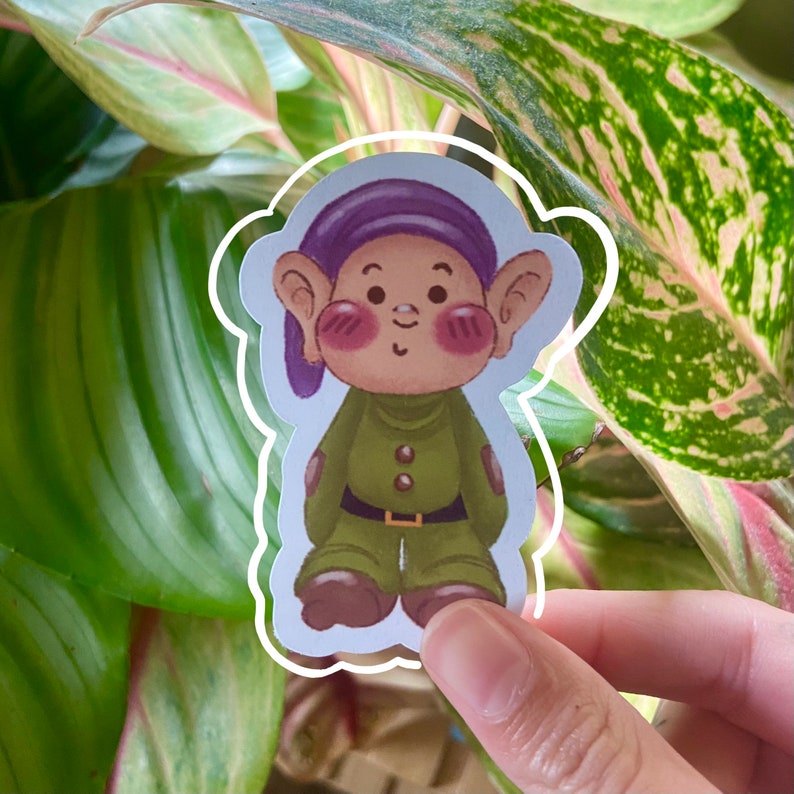 Puede incluir: Una pegatina de un enano de dibujos animados con un sombrero morado, una camisa verde y zapatos marrones. El enano tiene las mejillas sonrosadas y una expresi&oacute;n amigable.