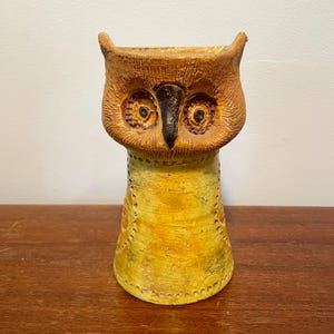 May include: A ceramic owl vase with a yellow and brown glaze. The owl's eyes are black and green, and the vase has a textured surface.