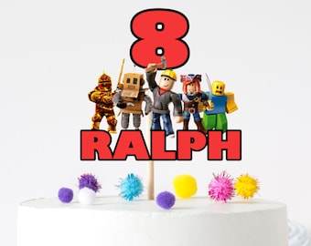 May include: A red and black cake topper with the number 8 and the name RALPH. The topper features a group of Roblox characters, including a knight, a robot, a builder, a pirate, and a yellow character. The topper is on a white cake with colorful pom-poms around the base.