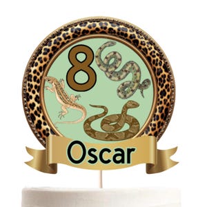 May include: A circular cake topper featuring a leopard print border, a light green centre with the number 8, a lizard, and two snakes. A gold banner at the bottom displays the name Oscar in black text. The topper is mounted on a thin stick.