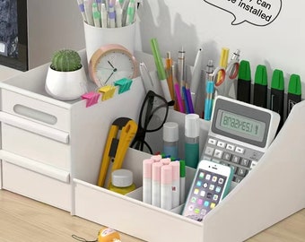 Puede incluir: Un organizador de escritorio de plástico blanco con dos cajones y múltiples compartimentos que contienen bolígrafos, una calculadora, un teléfono inteligente, un pequeño cactus en maceta, un reloj, tijeras y un cúter. El texto dice: Stationery can also be installed.