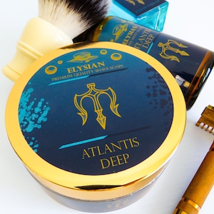 Può includere: Un contenitore di sapone da barba dorato e blu con un logo a tridente e il testo "Elysian Premium Quality Shave Soaps Atlantis Deep".