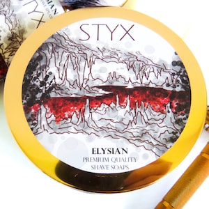 Puede incluir: Un recipiente redondo dorado con una etiqueta blanca que presenta una ilustración en blanco y negro de una cueva con agua roja. La etiqueta dice "Styx Elysian Premium Quality Shave Soaps".