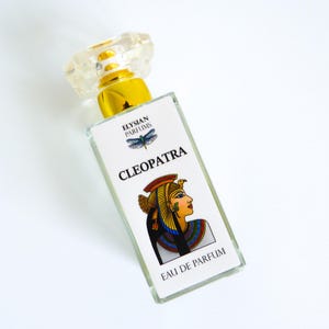 May include: A clear glass bottle of perfume with a gold cap and a white label. The label features a colourful illustration of Cleopatra and the text "Elysian Parfums Cleopatra Eau de Parfum".