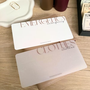 Può includere: Due carte rettangolari con testo. La carta in alto recita "EMERGENZA" in un font grande e stilizzato, con "emergenza" in una scrittura più piccola sotto. La carta in basso dice "VESTITI" con "vestiti" in corsivo. Entrambe le carte sono di colore chiaro.