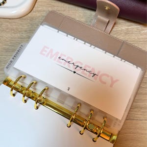 以下が含まれることがあります： ピンク色の「EMERGENCY」の文字が入った、ゴールドのリングが付いたベージュ色のプランナー。プランナーには「HIGH PRIORITY」と書かれたタブが付いています。プランナーは開いており、白いページが表示されています。