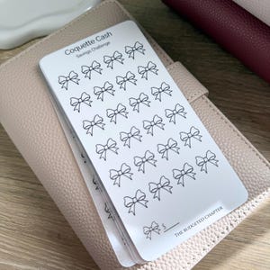 May include: White card with black bow illustrations, labeled "Coquette Cash Savings Challenge." The card is stacked on a light pink leather-like surface. The text "THE BUDGETED CHAPTER" is at the bottom of the card.