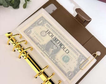 Könnte beinhalten: Ein brauner Ordner in Lederoptik mit einem goldenen Sechs-Ring-Mechanismus. Im Inneren befindet sich eine durchsichtige Kunststoffhülle mit einem Ein-Dollar-Schein und einer weißen Karte mit der Aufschrift HOUSEHOLD Cash Spending sowie eine goldene Büroklammer.