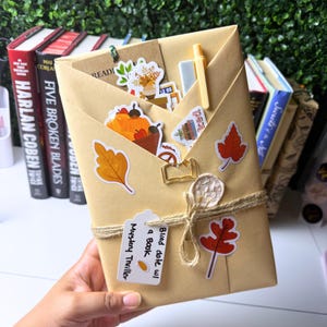 May include: A tan paper-wrapped book package decorated with autumn-themed stickers including pumpkins and leaves. It features a gold book-shaped paperclip, a twine tie with a wax seal, and a white tag labeled Blind date w/ a Book Mystery Thriller.