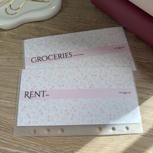 May include: Two cash envelope organizers with floral patterns and the words "GROCERIES" and "RENT" printed on them. The envelopes have a light pink band across the middle and are made of clear plastic with holes for a binder.