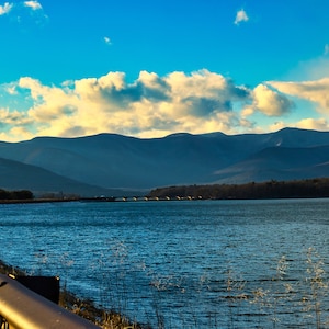 May include: A scenic view of a lake with a bridge in the distance, surrounded by mountains and a blue sky with white clouds.