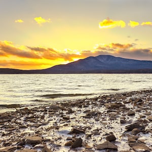 May include: A scenic view of a lake with a mountain range in the background. The sky is a vibrant orange and yellow, with clouds reflecting the sunset. The foreground shows a rocky shoreline.