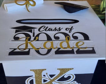 May include: A white graduation card box with black and gold lettering that reads "Class of 2022 Kade". The box has a black stripe and gold glitter lettering. A rolled diploma and graduation cap are on top.