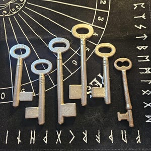 May include: Six antique metal keys of varying sizes and designs are arranged on a black surface with white markings. The keys have oval and figure-eight keyholes. Some keys have the numbers "4856" and "AE-429" stamped on them.