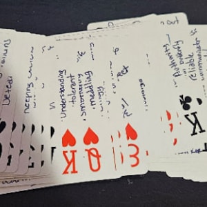 May include: A deck of playing cards with handwritten text on each card. The cards are arranged in a fan shape, with the top card showing a king of hearts and the text 'Understanding, tolerant, smiling,  meddling' and the bottom card showing a 7 of clubs and the text 'Unexpected'.