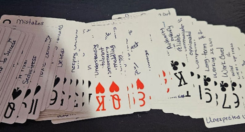May include: A deck of playing cards with handwritten text on each card. The cards are arranged in a fan shape, with the top card showing a king of hearts and the text 'Understanding, tolerant, smiling,  meddling' and the bottom card showing a 7 of clubs and the text 'Unexpected'.