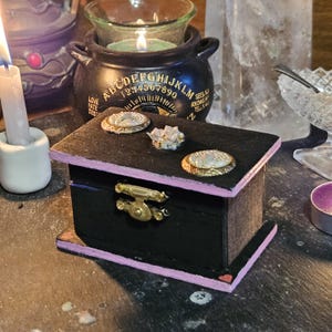 Puede incluir: Pequeña caja de madera negra con cierre dorado y ribete rosa. La parte superior de la caja presenta tres adornos decorativos. Rodeada de velas y otros objetos místicos, sugiere un tema de espiritualidad o adivinación.