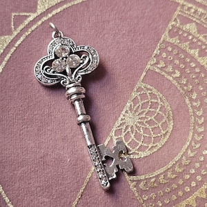 Puede incluir: Una llave de plata con una cabeza decorativa y adornada con un grupo de diamantes de imitación transparentes. La llave tiene un eje largo y delgado y un simple lazo en forma de corazón.