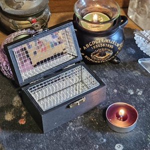 Könnte beinhalten: Eine offene schwarze Holzkiste mit verspiegeltem Innenraum und silbernem Drahtgitterdeckel. Eine kleine brennende Kerze und ein schwarzer kesselartiger Kerzenhalter mit einem Augensymbol und dem Alphabet sind ebenfalls vorhanden. Die Gegenstände sind auf einer dunklen, strukturierten Oberfläche angeordnet.