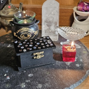 Puede incluir: Una caja de madera negra con una tapa decorada con piedras preciosas y un cierre dorado. Una pequeña vela roja arde cerca, y un caldero negro con el alfabeto y números está en el fondo. Un cristal de cuarzo transparente se alza.