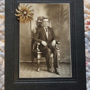 May include: A monochrome photograph of a man in a suit seated in a chair, set within a black frame. A gold-coloured brooch with pearl detailing is affixed to the frame. The photograph is likely from the early 20th century.