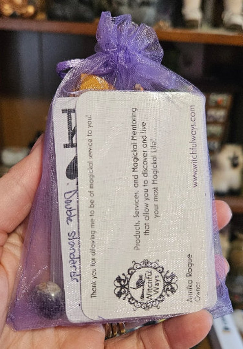 May include: A purple mesh bag with a white card inside. The card has the text 'Witchful Ways' and 'Products, Services, and Magickal Mentoring that allow you to discover and live your most Magickal Life!' The card also has the text 'www.witchfulways.com' and 'Annika Rogue Owner'.