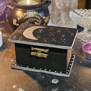 Puede incluir: Pequeña caja de madera negra con tapa con bisagras. La tapa está pintada con una luna creciente plateada y estrellas blancas, moradas y amarillas. La caja tiene un cierre dorado y está decorada con puntos blancos.
