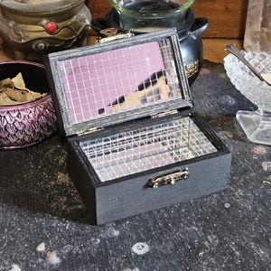 Könnte beinhalten: Kleine, offene schwarze Box mit verspiegeltem Innenraum und gitterförmigem Deckel. Die Box ist rechteckig mit einem Klappdeckel und einem kleinen Verschluss. Der Innenspiegel reflektiert einen rosafarbenen Farbton. Weitere Gegenstände im Hintergrund sind ein dekorativer Räuchergefäß und eine kleine Glasschale.