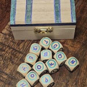 May include: A small wooden box with a hinged lid, painted in a light beige color with blue stripes. A collection of letter dice with blue lettering and borders are scattered in front of the box. The box has a gold-colored clasp.
