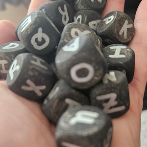 May include: A hand holding a pile of black wooden dice with white letters on each side. The letters are: R, O, N, K, H, X, O, A, I, and Z.