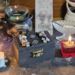 Puede incluir: Una caja de madera negra con un cierre dorado, adornada con piedras preciosas de colores y pequeños amuletos en forma de casa. Una pequeña vela roja arde cerca, y una olla decorativa con una vela está en el fondo. La olla tiene letras y números.