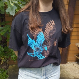 Op de afbeelding: Een zwart T-shirt met een blauwe en bruine adelaar-grafische print. De adelaar is in vlucht met zijn vleugels wijd uitgespreid.