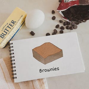 Könnte beinhalten: Ein Stillleben mit Backzutaten. Ein Stück gesalzene Butter, ein Ei und eine Tüte Schokoladenstückchen sind um einen Notizblock mit einer Zeichnung eines Brownies und dem Wort "Brownies" angeordnet.